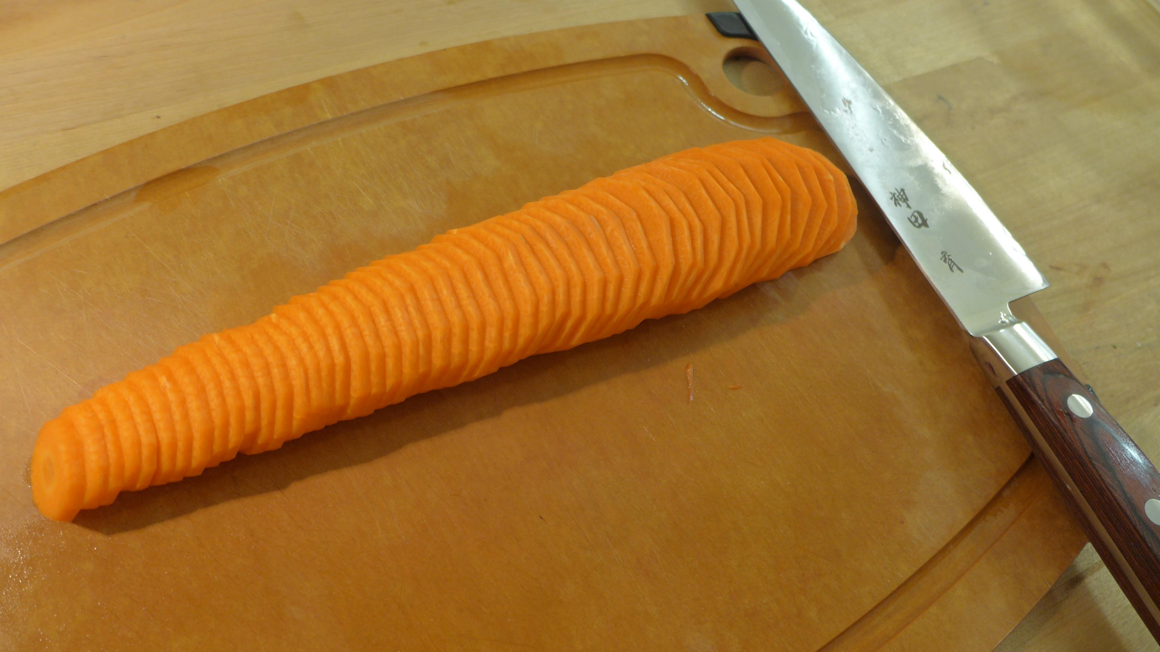 砧板上有一根紅蘿蔔，整支被切成片，紅蘿蔔片彼此側疊著。旁邊有一支上面寫著「神田 育」的牛刀