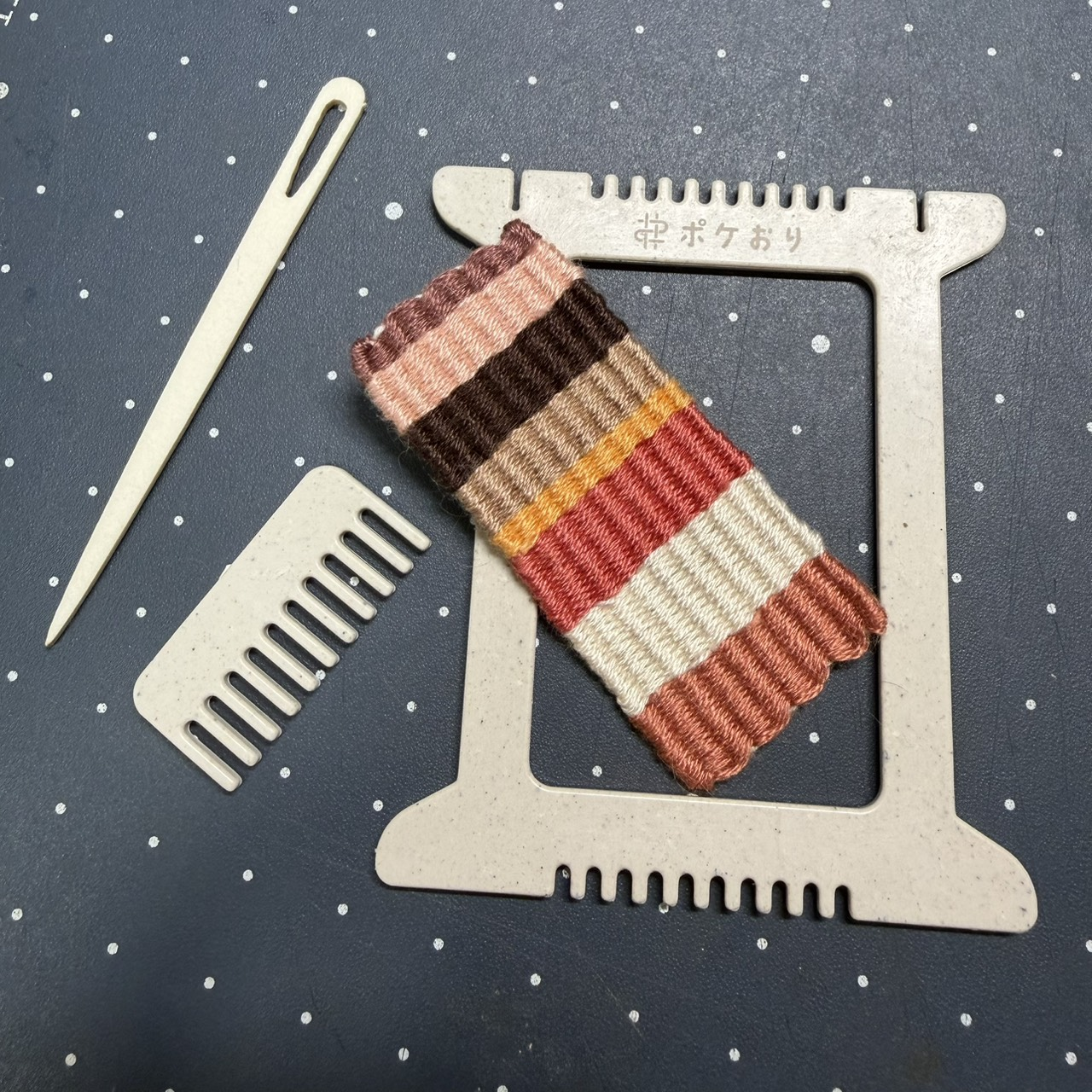 一片有鋸齒狀凹槽的織板和梳子狀的配件和針，上面放著一小塊織好的條紋布