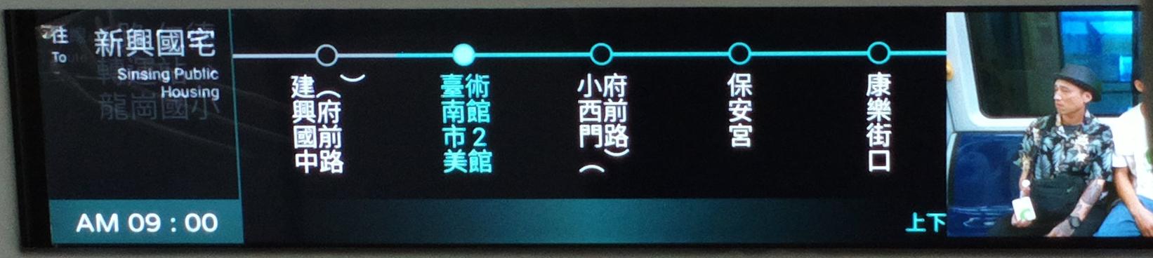 公車車廂內照片擷取行駛資訊看板區域,包含左側五分之一區塊、中央五分之三區塊、右側五分之二區塊。左側區塊主要內容為黑底白字橫書,文字正從「路線 6 路 仁德轉運站→龍岡國小」變換為「往 新興國宅」,僅「路線」、「往」、「新興國宅」底下並陳英文;此區塊文字對右靠齊。區塊底端以綠底白字顯示「AM 09:00」。中央區塊以線條標示部分停靠站,由左至右分別為前一站、下一站、下下站、下下下站、下下下下站,已過路線以白色線條標示、未抵達路線以綠色線條標示,各站點以空心圓形標記,僅下一站站點以實心圓形標記;各站名稱以直書由左至右方向書寫,縱向顯示文字字數固定為四字,包含全型上下括號及阿拉伯數字,未避頭尾,僅下一站站名文字為綠色,前後其餘各站站名文字均為白色。區塊底端採用黑綠黑漸層底色顯示綠色文字跑馬燈。右側區塊持續播放廣告宣導影片。