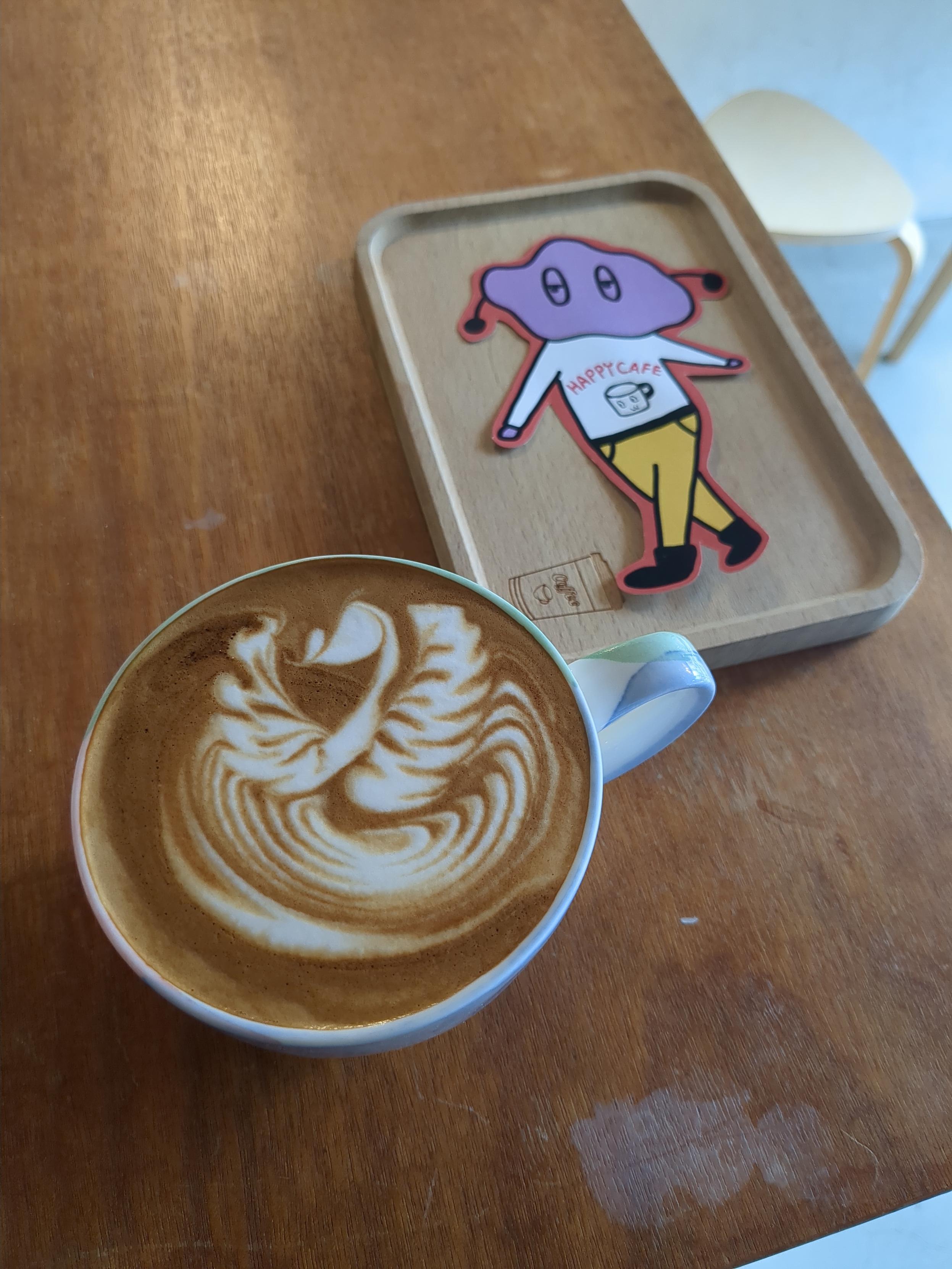 一杯有雙翅鵝拉花的拿鐵，旁邊的木托盤上有一個插畫人物，頭部像一朵雲，身上的衣服寫著HAPPY CAFE，呈現在正在走路的樣子。